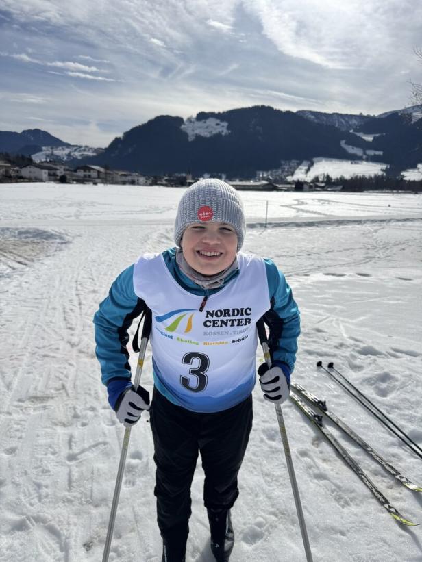 Ein Kind mit Startnummer 3 vom Nordic Center steht lächelnd mit Skiern in einer verschneiten Landschaft.