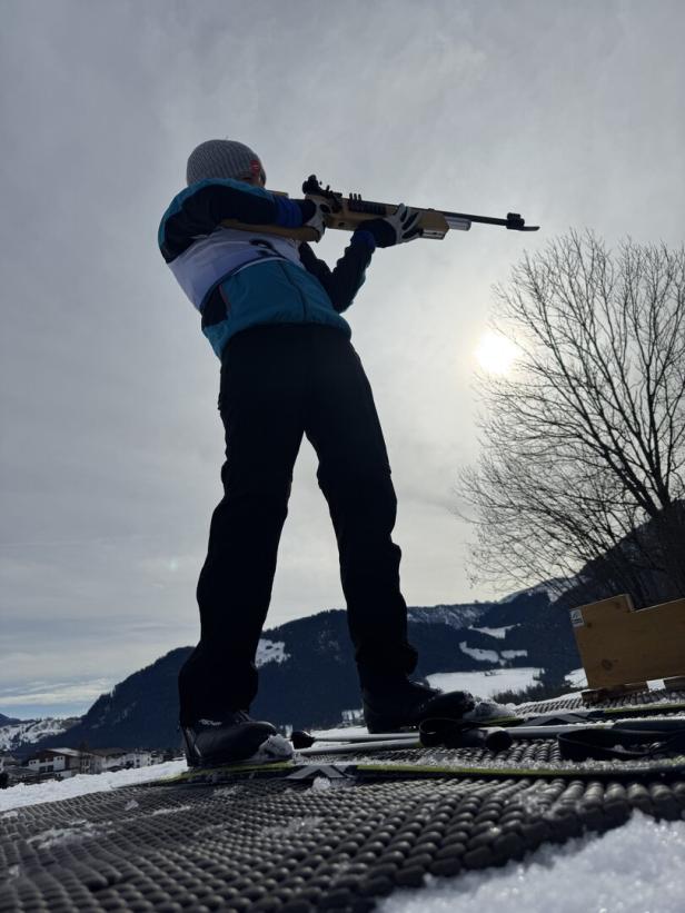 Ein Biathlet zielt mit einem Gewehr vor einer verschneiten Berglandschaft in den Himmel.