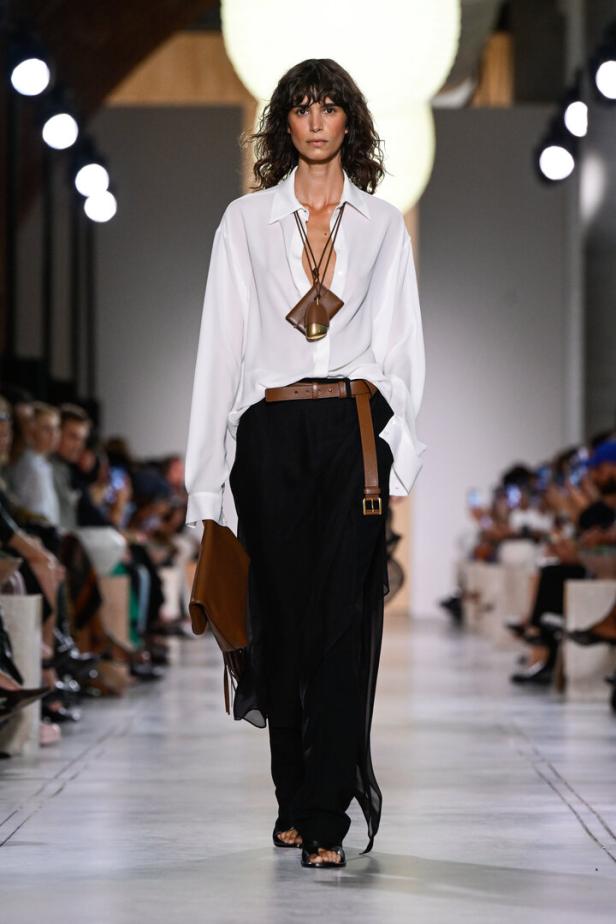 Frau am Laufsteg in weißer Bluse, braunem Gürtel und schwarzer Hose