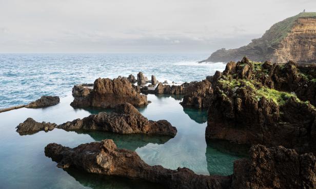 Felsenbecken mit klarem Wasser vor einer Küstenlandschaft unter bewölktem Himmel.