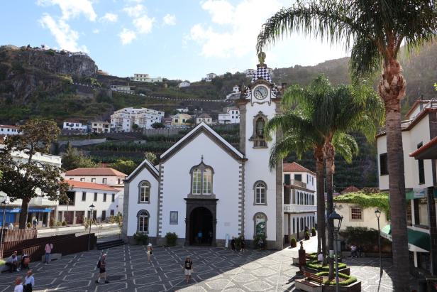 Eine Kirche mit Uhr steht auf einem Platz, im Hintergrund ein bewaldeter Hügel mit Häusern.