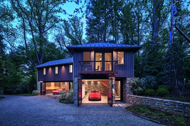 Modernes Haus mit großen Fenstern und zwei offenen Garagen, in denen rote Sportwagen stehen, umgeben von Bäumen und Garten.