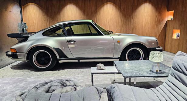 Ein silberner Oldtimer-Sportwagen steht in einem modern eingerichteten Raum mit Holzwand und Designer-Möbeln.