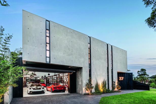 Modernes Haus mit klaren Linien, großen Fenstern und einer offenen Garage, in der zwei Sportwagen stehen.