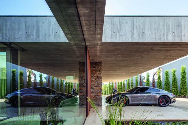 Ein grauer Sportwagen steht unter einem modernen Carport mit Beton- und Glaselementen, umgeben von hohen, grünen Pflanzen.