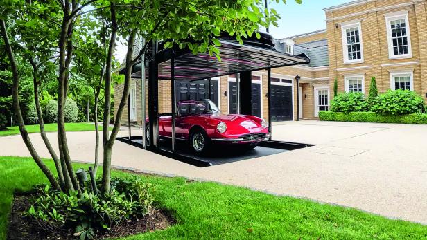 Ein rotes Cabrio steht auf einer versenkbaren Auto-Plattform vor einem eleganten Haus mit gepflegtem Garten.