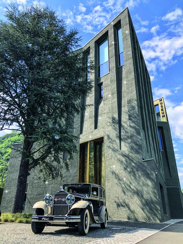 Ein Oldtimer steht vor einem modernen, hohen Museumsgebäude mit großen Fenstern und einem Baum daneben.