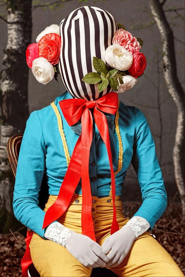 Eine Person mit gestreifter Kopfbedeckung, Blumen und Blättern am Kopf trägt ein farbenfrohes Kostüm mit roter Schleife.