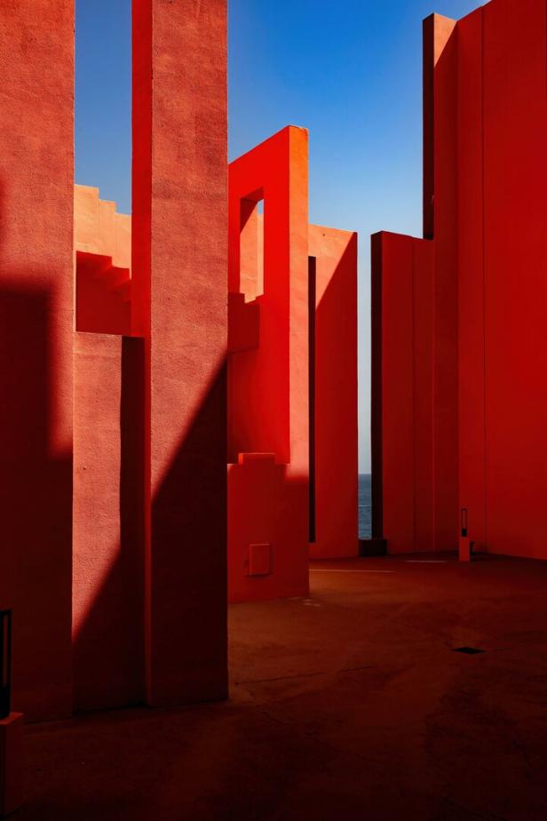 Rote, geometrische Gebäudestrukturen mit Schatten und Blick auf das Meer unter klarem, blauem Himmel.