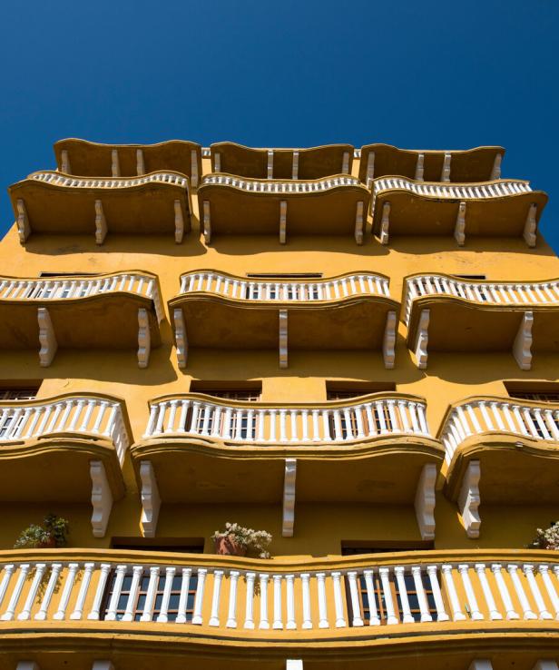 Gelbes Gebäude mit mehreren Stockwerken und weißen, geschwungenen Balkongeländern vor blauem Himmel.