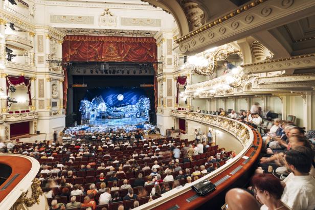 Zuschauer sitzen in einem prunkvollen Opernhaus und warten auf den Beginn einer Aufführung.