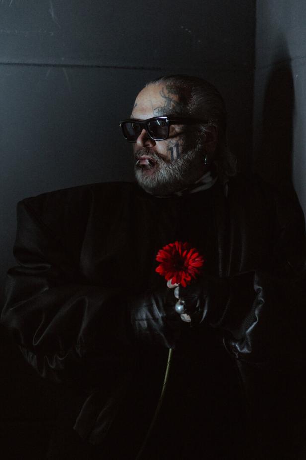 Person mit Sonnenbrille, Gesichtstattoos und Handschuhen hält eine rote Blume in dunkler Umgebung.