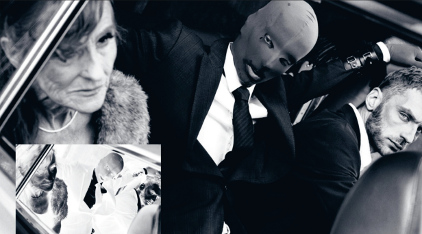 Eine ernst dreinblickende Frau sitzt neben zwei Männern, beide in Anzug. Einer hat eine Sturmhaube auf.