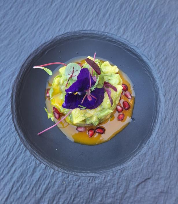 Auf einem dunklen Teller befindet sich ein Avocado-Tatar mit Granatapfelkernen und einer lila Blüte.