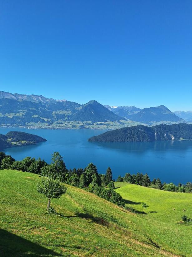 Grüne Hügel führen zu einem blauen See, dahinter erheben sich Berge unter strahlend blauem Himmel.