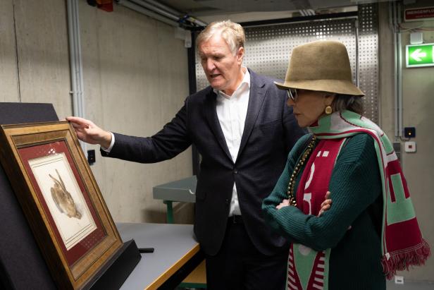 Ein Mann zeigt einer Frau mit Hut und Schal ein gerahmtes Bild eines Hasen in einem Raum mit Betonwänden.