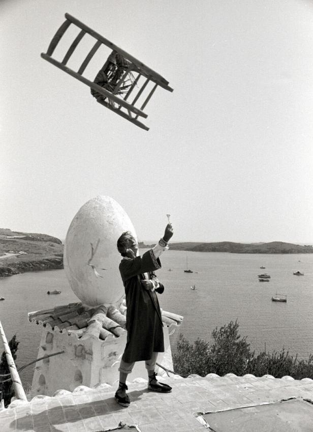 Ein Mann steht auf einer Terrasse am Meer, hält einen kleinen Gegenstand hoch, während ein Stuhl durch die Luft fliegt.