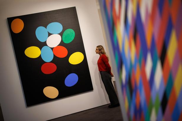 Eine Frau betrachtet ein Gemälde mit bunten ovalen Formen auf schwarzem Hintergrund, im Vordergrund unscharfe bunte Muster.