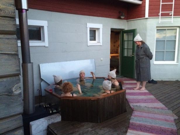 Fünf Personen mit Mützen entspannen in einem Außen-Whirlpool, eine weitere Person steht im Bademantel daneben.