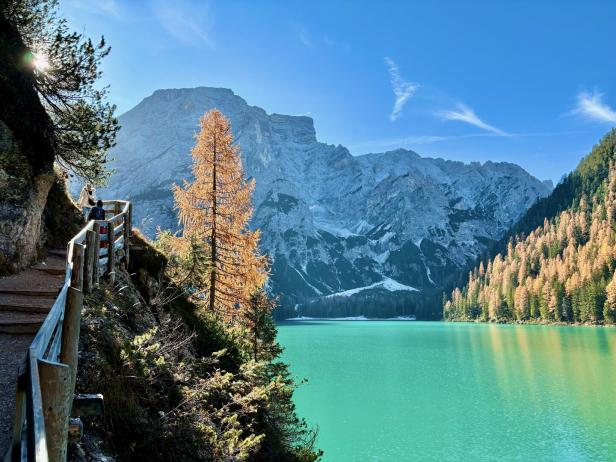Ein Bergsee liegt vor einer Bergkette, flankiert von einem bewaldeten Ufer und einem Weg mit Wanderern.