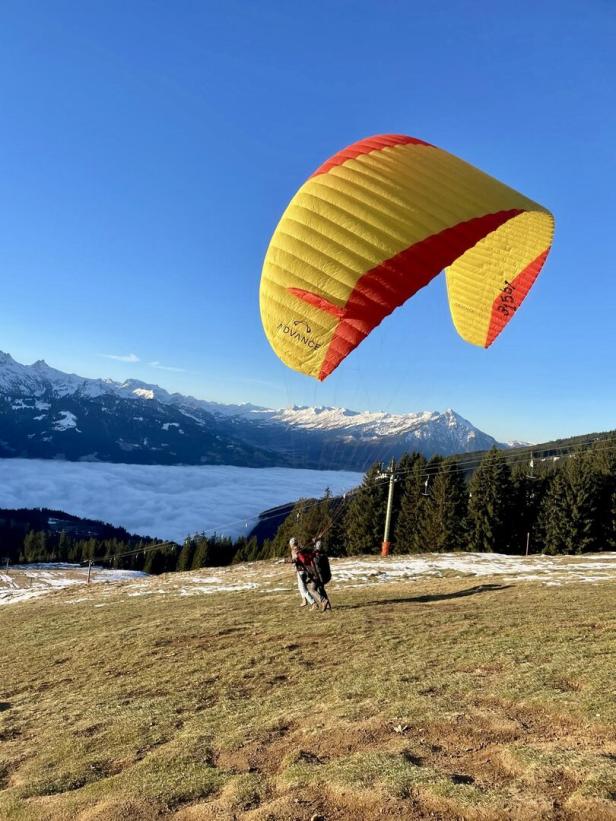 Ein gelb-roter Gleitschirm schwebt über einer Person auf einem Hügel mit Blick auf verschneite Berge.