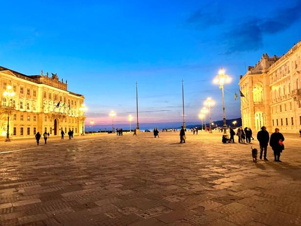 Ein Platz mit Menschen und beleuchteten Gebäuden unter blauem Himmel in Triest.