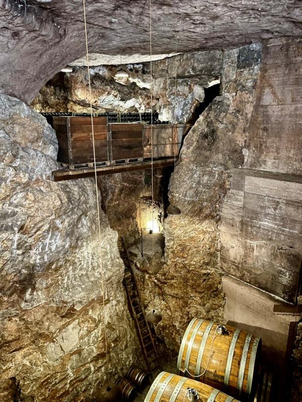 In einem Felsenkeller stehen Holzfässer und eine Leiter unter einer Holzbrücke mit Kisten.