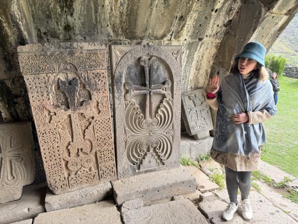 Eine Frau steht neben reich verzierten Steintafeln mit eingravierten Kreuzen und erklärt etwas mit erhobener Hand.