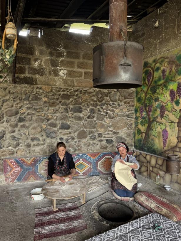 Zwei Frauen bereiten auf bunten Kissen sitzend Teig vor einem traditionellen Ofen in einem rustikalen Raum mit Steinwänden zu.