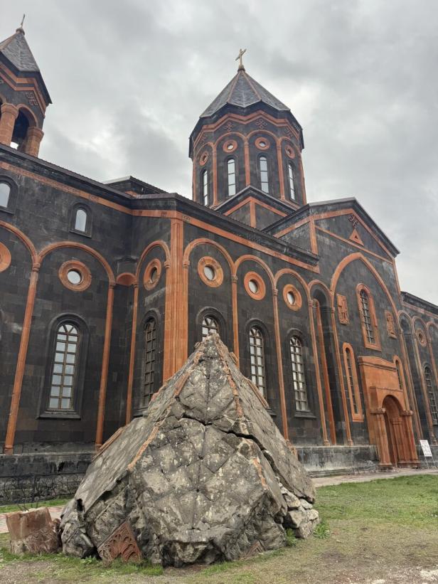 Dunkle Kirche mit orangefarbenen Verzierungen, davor ein großer, zerbrochener, verzierter Stein auf einer Wiese.