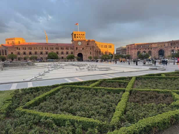 Ein großer Platz mit gepflegten Grünflächen, umgeben von historischen Gebäuden aus rötlichem Stein und vielen Menschen.