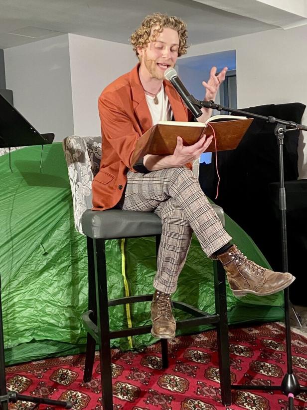 Ein Mann mit lockigem Haar liest aus einem Buch in ein Mikrofon, sitzend auf einem Stuhl.