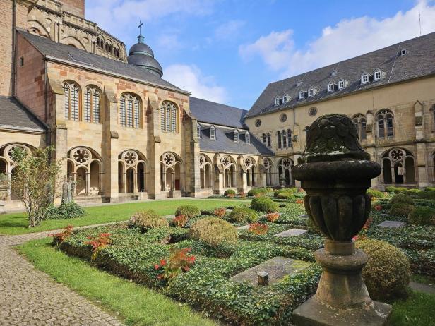 Ein Innenhof mit Rasenflächen, Blumenbeeten und alten Gebäuden mit Arkaden unter blauem Himmel.