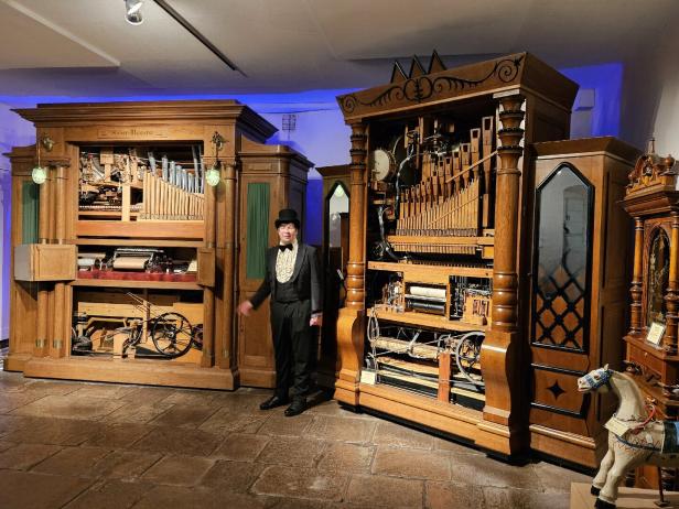 Ein Mann im Anzug steht vor zwei großen, mechanischen Musikinstrumenten in einem Raum.