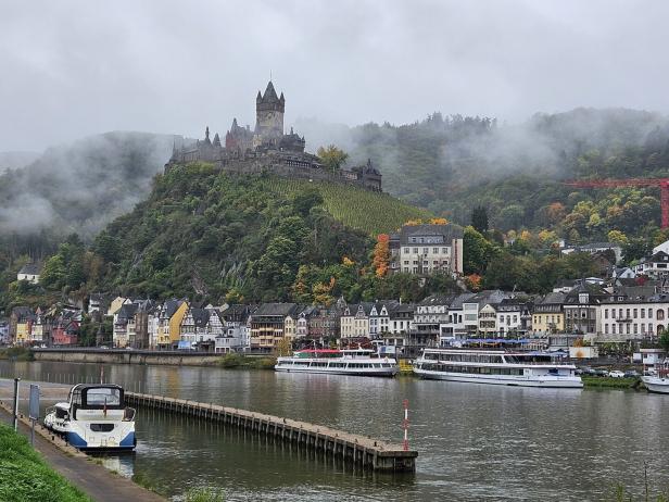 Eine Burg thront auf einem bewaldeten Hügel über einer Stadt am Flussufer.