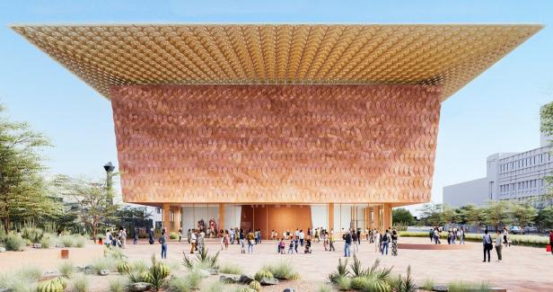 Ein modernes Gebäude mit großer, goldener Überdachung, kupferfarbener Fassade und vielen Menschen davor, umgeben von Wüstenpflanzen.