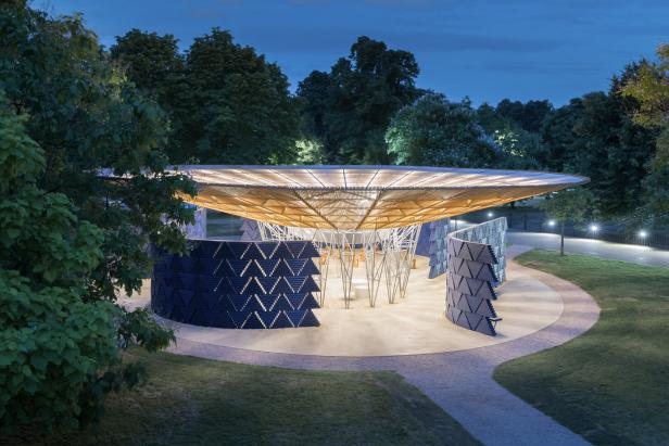 Pavillon mit großem, beleuchtetem Dach aus Holz und Metall, umgeben von geometrisch gemusterten Wänden im Park bei Dämmerung.