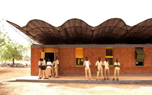 Mehrere Jugendliche in Schuluniform stehen vor einem Backsteingebäude mit wellenförmigem Dach und gelben Fensterläden.