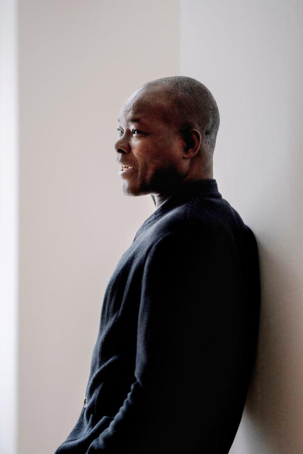 Ein Mann mit kurzem Haar lehnt seitlich an einer hellen Wand und blickt nach vorne.