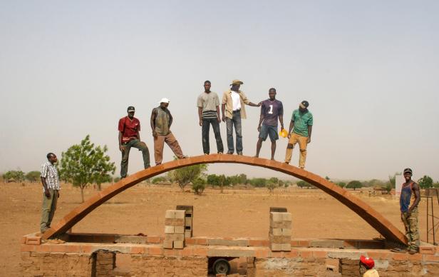 Sieben Männer stehen auf einem gemauerten Rundbogen, zwei weitere stehen daneben, im Hintergrund trockene Landschaft mit Bäumen.