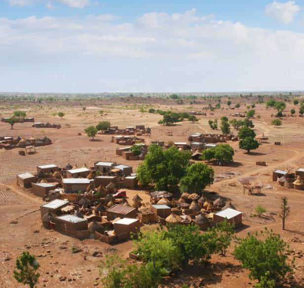Mehrere traditionelle Lehmhäuser mit Strohdächern und einige grüne Bäume in einer trockenen, weitläufigen Landschaft.