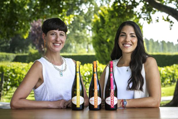 Zwei Frauen sitzen an einem Holztisch im Freien mit drei Flaschen Chandon Spritz vor sich und lächeln in die Kamera.