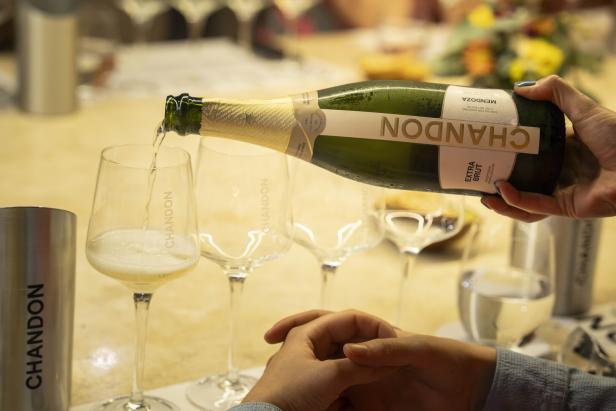 Eine Hand schenkt Schaumwein aus einer Chandon-Flasche in ein Glas, weitere Gläser und Dekoration im Hintergrund.