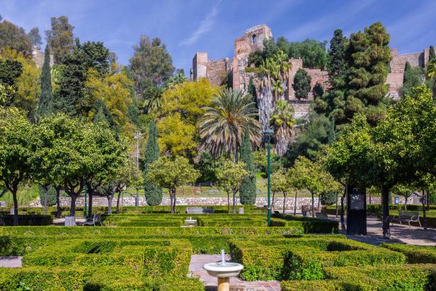 Malaga Spain lush greenery and architectural fountains in the Pedro Luis Alonso Gardens with the historic stone walls of the Alcazaba of Malaga fortress in the background.