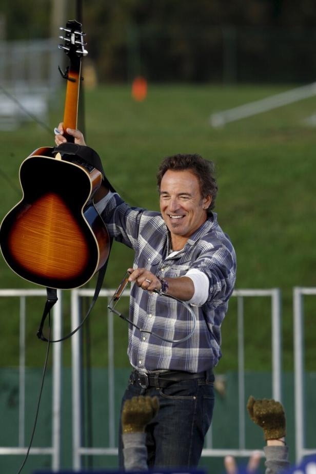 Bruce Springsteen hält lächelnd eine Gitarre hoch und trägt ein kariertes Hemd.