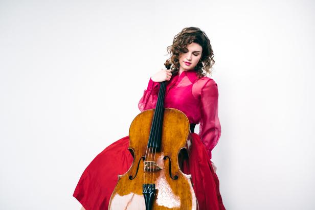 Frau mit lockigen Haaren in rotem Kleid hält ein Cello vor weißem Hintergrund.