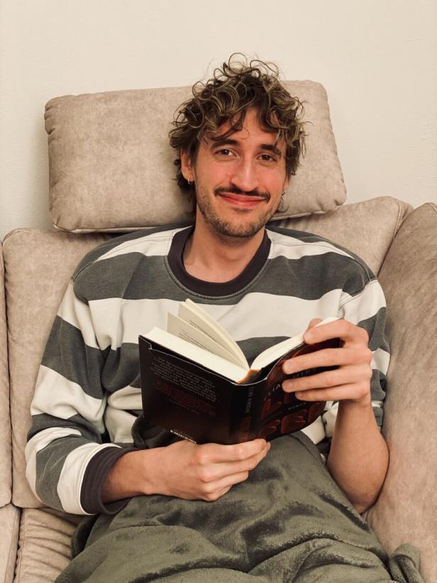 Ein junger Mann sitzt entspannt auf einem Sofa, liest ein Buch und lächelt, während er in eine Decke gehüllt ist.