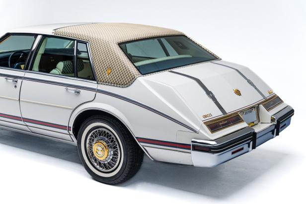 Luxuriöses Auto mit beigem Gucci-Muster am Dach, goldenen Details und auffälligen Felgen, weißer Lackierung und roten Zierstreifen.