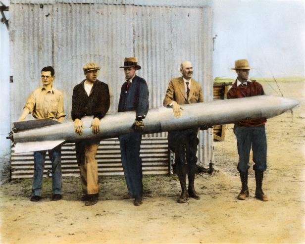 Fünf Männer stehen nebeneinander und halten gemeinsam eine große silberne Rakete vor einem Blechschuppen.