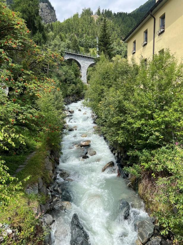 Ein Gebirgsbach fließt zwischen grünen Bäumen und einem gelben Haus, im Hintergrund eine Steinbrücke und bewaldete Hügel.